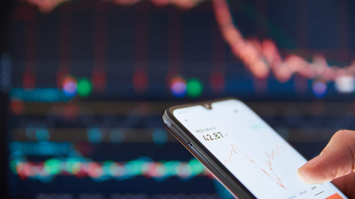 Safe Money Moves People Rely On Now Person checking the stock prices on a smartphone in front of a big scre